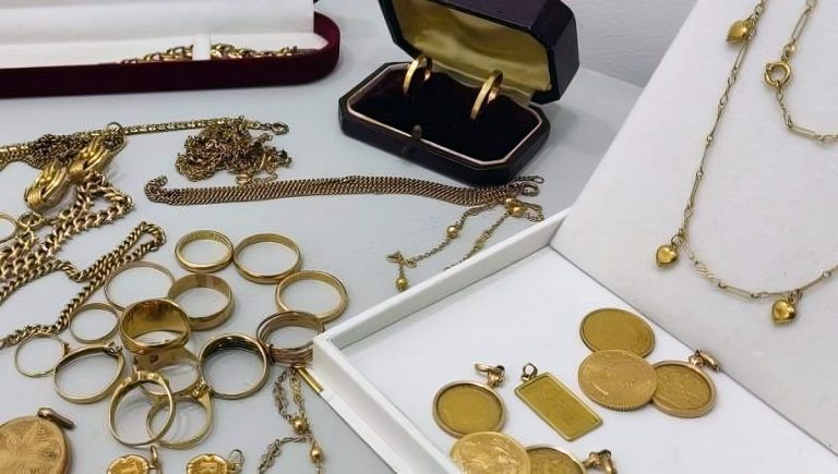 Imagem com diferentes peças em ouro, moedas, barras, fios, pulseiras, aneis, medalhas, brincos, dispostos alguns dentro de caixas de joalharia outras peças dispostas sobre mesa.